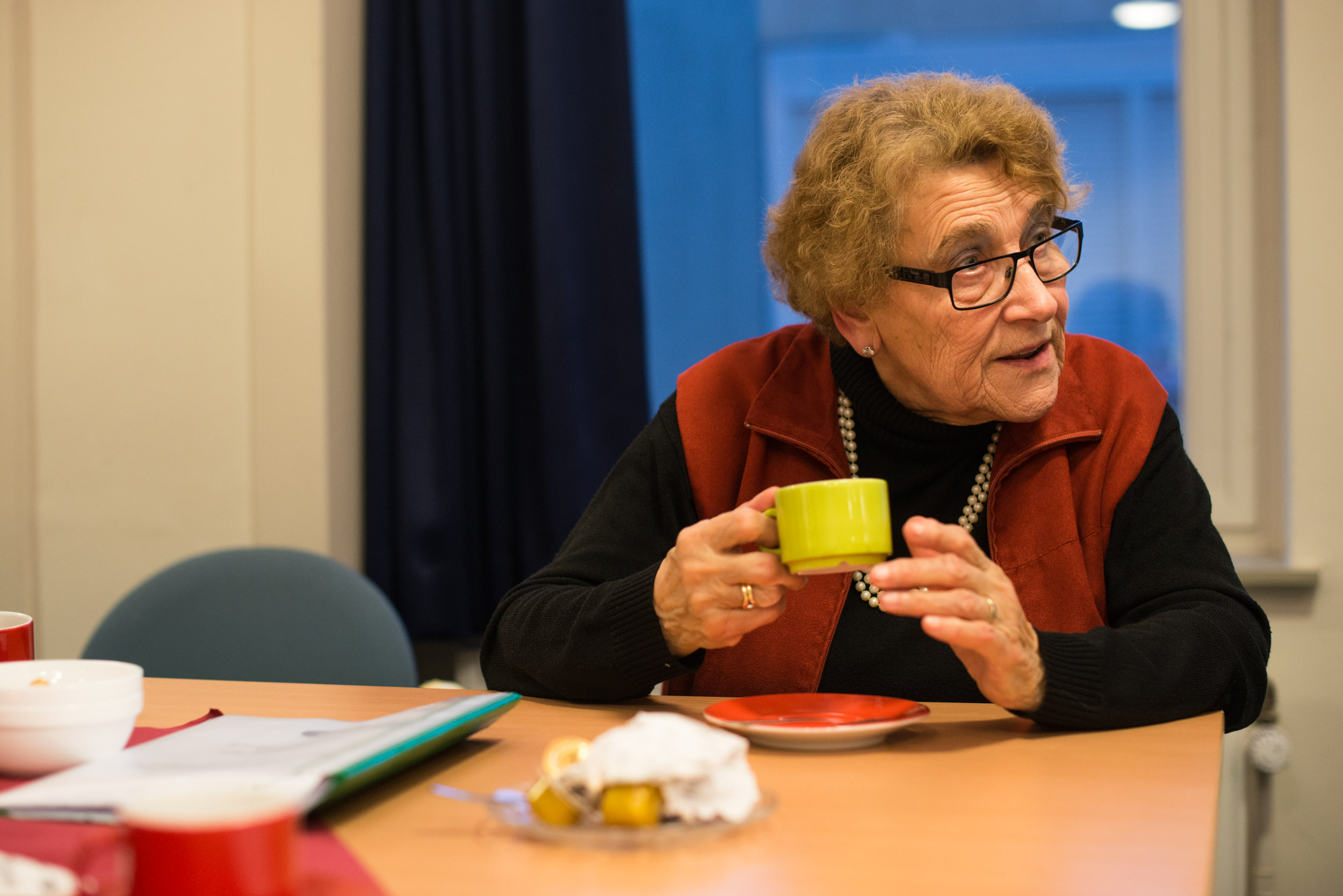 oudere vrouw met grijs haar, zwarte bril en een gele mok in haar handen