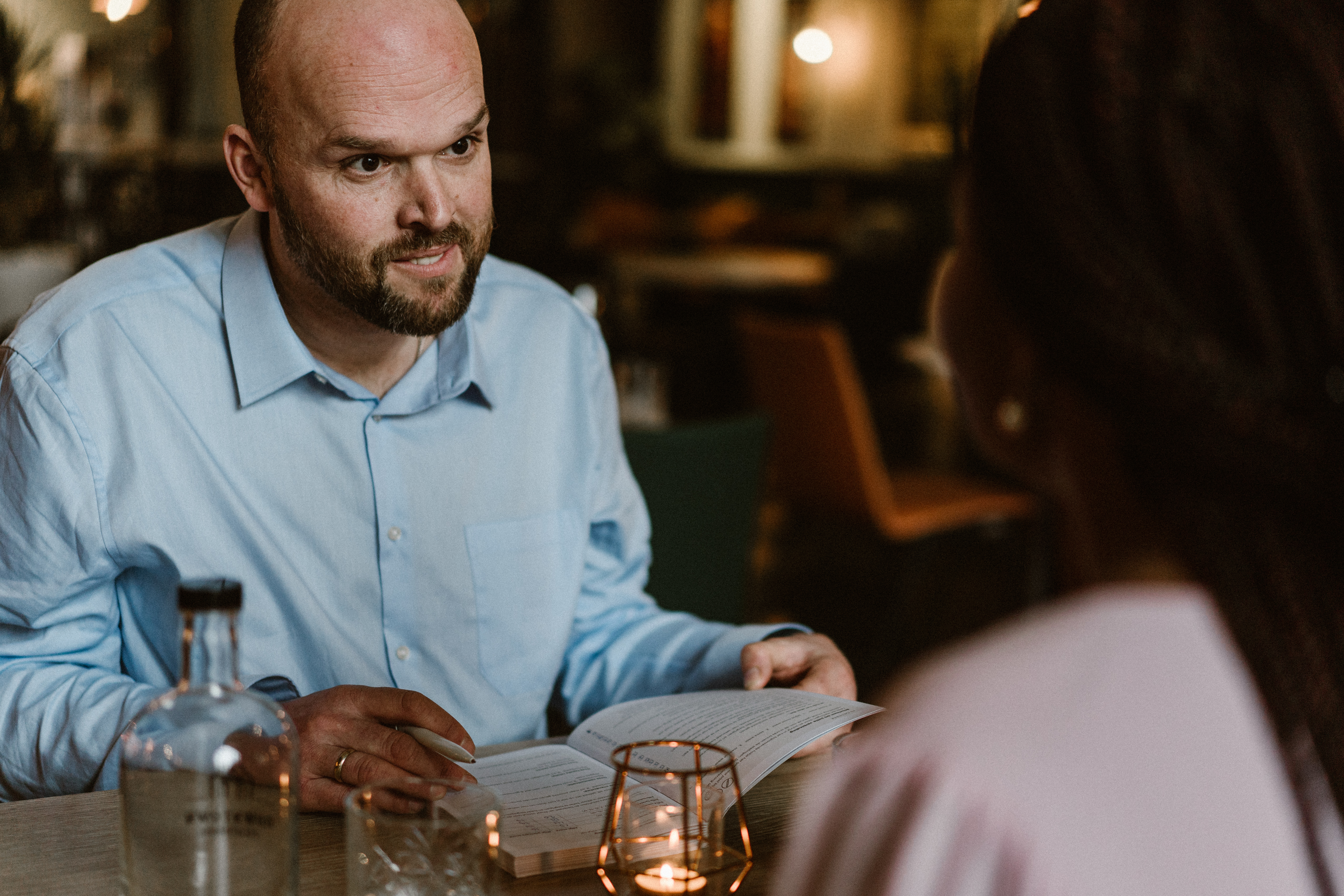 Man, kalend, met kort getrimde baart, zit tegenover een vrouw die half zichtbaar is vanaf de rug. Man heeft een boek opengeslagen voor zich