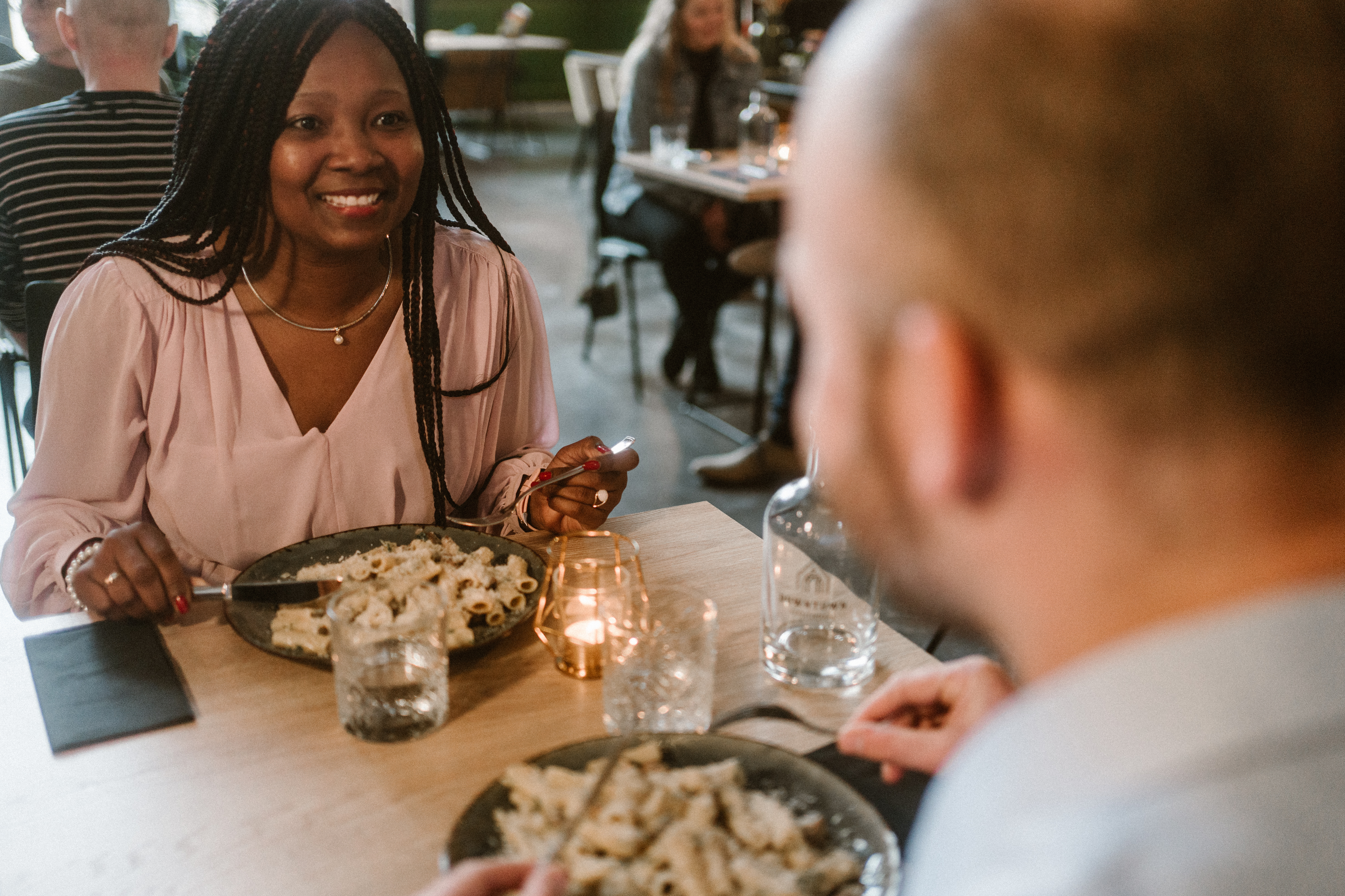 Man en vrouw die aan 't eten zijn, voor hun een bord pasta. Man is vaag zichtbaar en vanaf de rug. Vrouw heeft een glimlach.